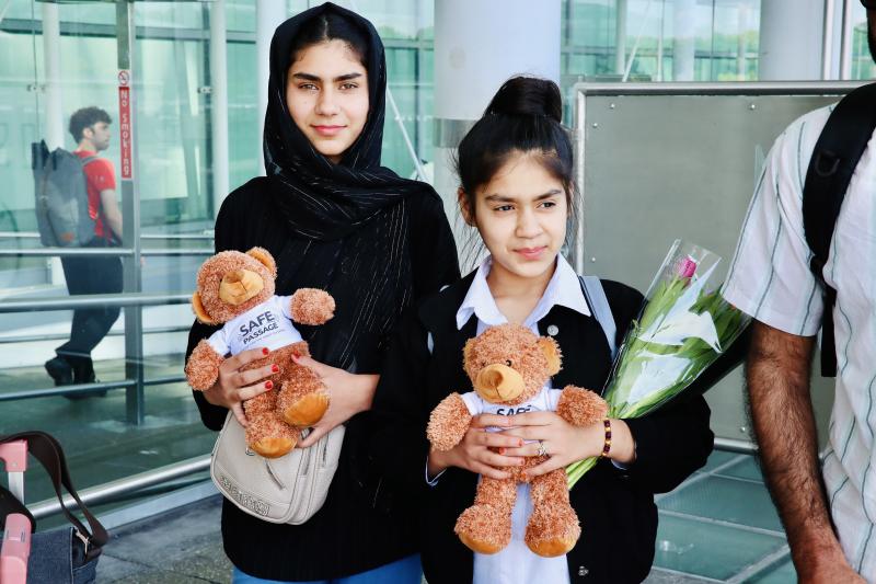 Picture of two young girls standing in front of a UK airport, with 'Safe Passage' branded teddy bears in their arms 